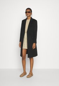 Palton lung negru cu un design croit și două butoane, asociat cu un top bej și o fustă texturată aurie, completat cu pantofi slip-on de culoare maro deschis.