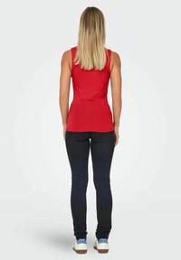 Vrouw met blond haar die met haar rug naar de camera staat, gekleed in een rode mouwloze top, zwarte skinny jeans en witte sneakers met blauwe accenten.