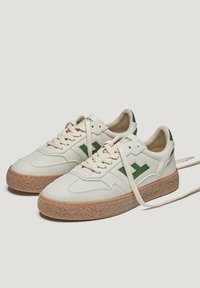 Baskets en cuir blanc avec accents verts, bout rond, design à lacets, semelle en caoutchouc crème texturée et détail du logo sur le côté.