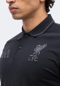Homme portant un polo noir du Liverpool FC avec écusson du club brodé et logo Adidas sur la poitrine, col boutonné.