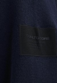Tessuto di un blu navy con una prominente etichetta nera a toppa che presenta i dettagli del marchio, inclusa la dicitura "UNLTD CORE" e informazioni sulle taglie.