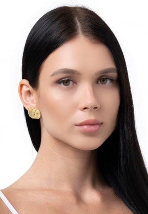 Goldfarbene, strukturierte Ohrringe mit einem welligen, überlappenden Design. Ein Modell mit langen, glatten, dunklen Haaren posiert vor einem weißen Hintergrund.