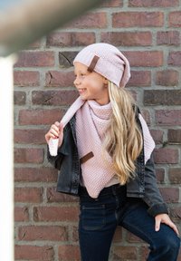 Różowy dzianinowy czapka i szalik z teksturowymi detalami, w połączeniu z czarną dżinsową kurtką i niebieskimi dżinsami, na tle ceglanej ściany.