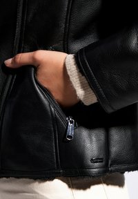 Czarna skórzana kurtka z boczną kieszenią na zamek, charakteryzująca się teksturowanym wykończeniem. Rękawy mają delikatny beżowy detal z tkaniny.