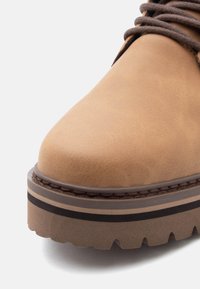 Close-up of a tan leather boot with thick, multi-layered sole and brown laces on a white background.