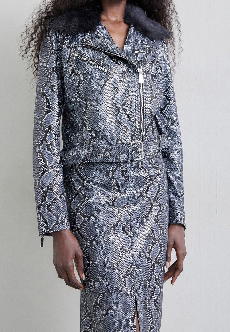 Femme portant une veste grise et noire à motif peau de serpent avec ceinture et une jupe assortie fendue à l'avant, debout contre un mur uni.