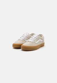 Beige velúr sportcipők fehér fűzőkkel, oldalsó fehér swoosh jellel és gumírozott talppal. Lekerekített orr és varrott részletek az anyagon.