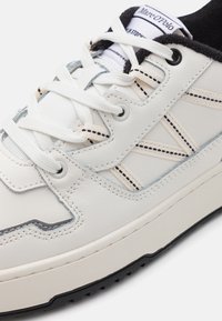 Hvid lædersneaker med tekstureret sort gummisål. Har lysfarvet syning, meshlæg og subtilt mønstrede detaljer.