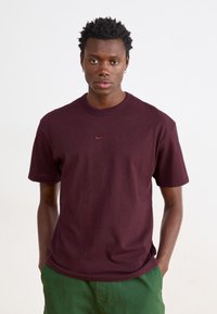 Bordeaux-koton T-paita lyhyillä hihoilla, pyöreä kaula-aukko ja pieni, kohokuvioitu Nike-logo rintakehässä, yhdistettynä vihreisiin shortseihin.