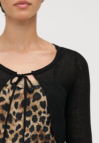 Crna, pletena cardigan s okruglim izrezom i zatvaranjem na mašnu sprijeda. Nosena preko majice s leopard uzorkom i teksturiranom površinom.