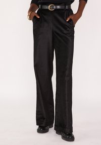 Pantalon large en satin noir à la texture lisse, avec poches latérales et ceinture plate. Porté avec une ceinture noire et des chaussures noires.