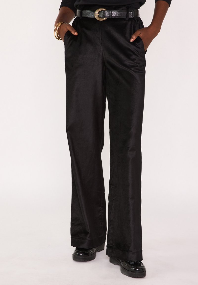 Pantalon large en satin noir à la texture lisse, avec poches latérales et ceinture plate. Porté avec une ceinture noire et des chaussures noires.