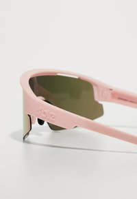 Bliz saulesbrilles ar mīksta rozā plastmasas rāmi un zaļgandīgtas lēcas. Tam ir elegants dizains un redzams logotips uz tempļa.