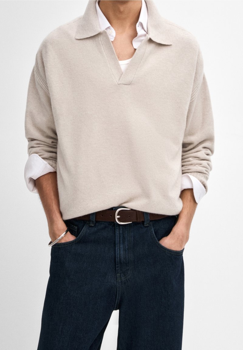 Pull-over beige en maille avec un col en V et un col plié, porté sur une chemise blanche. Associé à un jean bleu foncé et une ceinture marron.