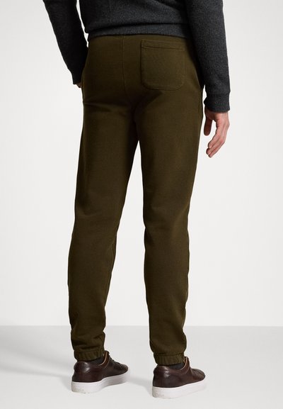 Oliven grønne joggebukser med innsnevrede anklene, elastisk midjebånd, enkelt baklomme, laget av mykt stoff, brukt med mørke sko.