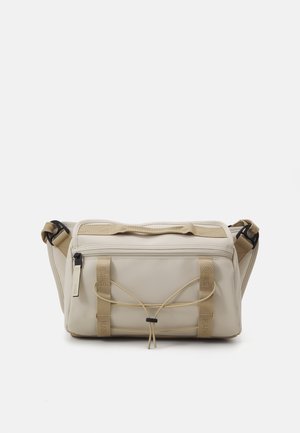 Sac bandoulière rectangulaire beige avec poche zippée à l'avant, sangle réglable et détail en cordon élastique sur surface lisse.