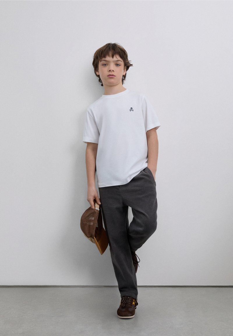 T-shirt blanc à manches courtes avec un petit logo noir, associé à un pantalon gris foncé et des chaussures marron. Une casquette en velours côtelé marron est tenue dans une main.