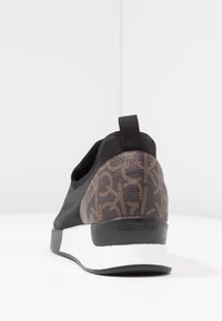 Czarny but wsuwany z teksturowaną brązową tylnią częścią z motywem logo, białą podeszwą środkową i czarną gumową podeszwą zewnętrzną.