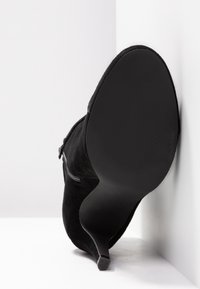 Bottine en daim noire à talon haut vue de la semelle, posée contre un mur d'angle blanc sur une surface blanche.