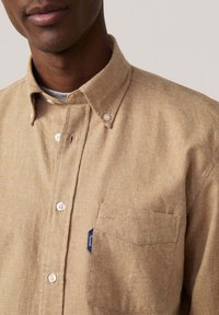 Camisa de algodón color marrón claro con cuello abotonado, botones frontales, bolsillo parche y pequeña etiqueta de la marca. Textura suave con un sutil patrón jaspeado.