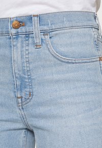 Jeans de ganga azul claro com cintura média, apresentando um botão de cobre, detalhes de costura subtil e um design padrão de cinco bolsos.