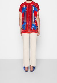 Haut à manches courtes à motifs rouges avec des motifs d'oiseaux bleus et de fleurs, associé à un pantalon large crème et des sandales bleues.