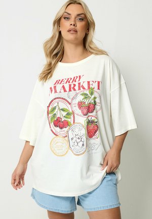 Kvinde iført en oversize hvid T-shirt med rød "Berry Market" tekst og farverige bærillustrationer, kombineret med lyseblå denimshorts.