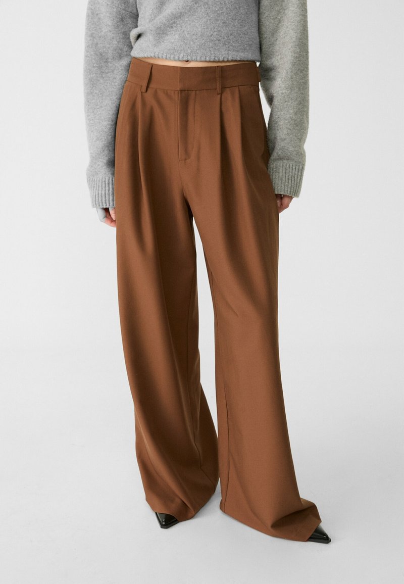 Hnědé široké kalhoty vyrobené z hladké tkaniny, s plisováním v přední části, vysokým pasem a upraveným střihem.
