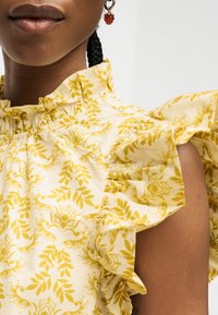 Gros plan d'une personne portant un chemisier jaune à volants et fleurs ainsi qu'une boucle d'oreille rouge perlée, avec des cheveux tressés visibles.