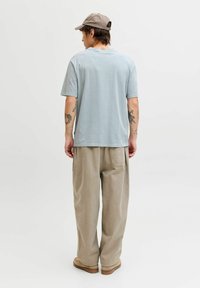 Hellblaues Baumwoll-T-Shirt mit kurzen Ärmeln und Rundhalsausschnitt, kombiniert mit beigen Hosen mit weitem Bein. Der Look wird durch dezente Tattoos und eine Kappe ergänzt.