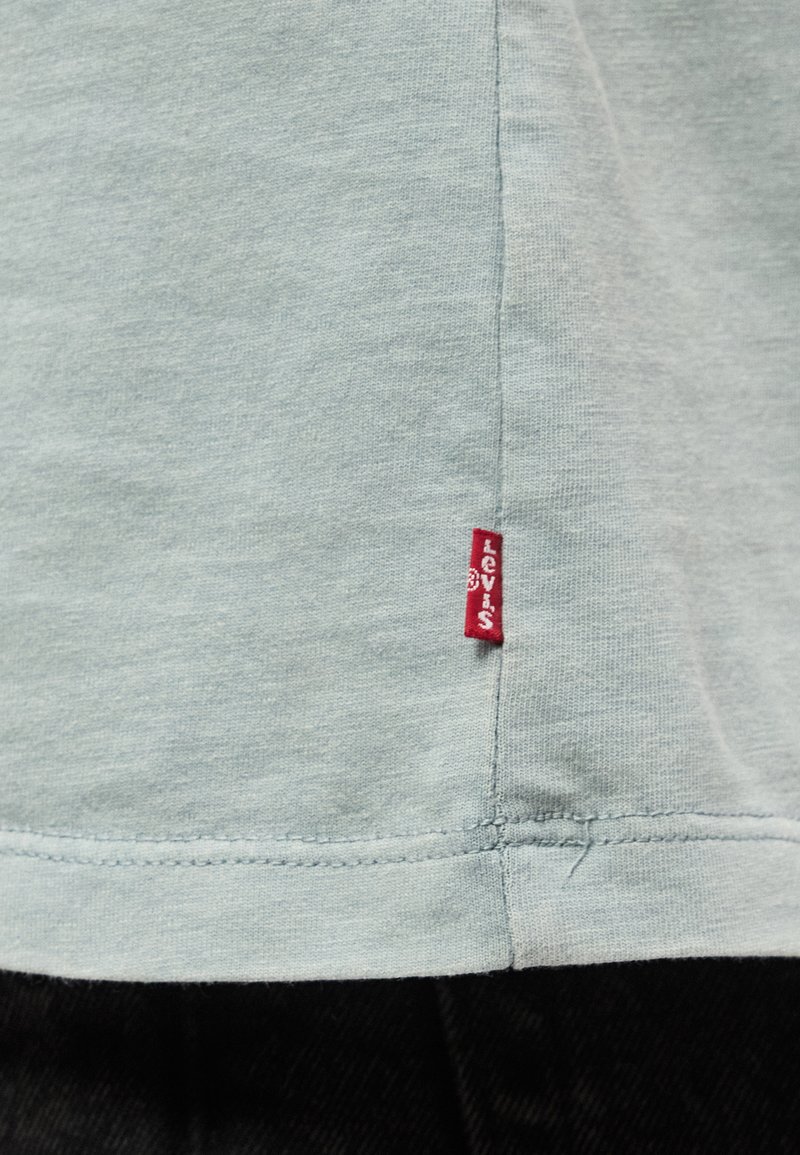 Tela gris clara con dobladillo de doble costura y una pequeña etiqueta vertical roja de Levi's cosida cerca de la costura sobre tela negra.