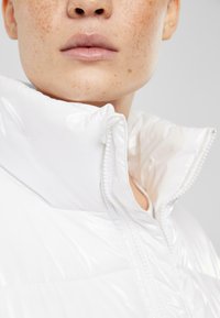 Hvit, glinsende pufferjakke med høy krage, som har intrikate sømmer og glidelås. Den glatte teksturen fremhever det lette designet.
