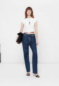 Witte cropped t-shirt met grafische print, blauwe rechte jeans, zwarte handtas en zwarte sandalen met hakken op een effen witte achtergrond.