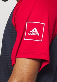 T-shirt de sport Adidas rouge et marine avec manches courtes, motif texturé et patch logo blanc sur l'épaule gauche.