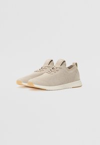 Beige Strick-Sneaker mit strukturierter Oberseite, abgerundetem Zehenbereich und kontrastierender, hellbrauner Gummisohle. Verfügt über Schnürverschlüsse und Logoakzente.