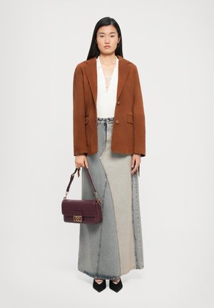 Woman stands wearing a rust brown blazer, white lace-up top, two-tone denim maxi skirt, black pointed shoes, and holding a dark purple handbag.