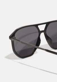 Lunettes de soleil noires avec une monture géométrique, verres sombres et accents en métal. Les branches présentent un design élégant avec le nom de la marque gravé.