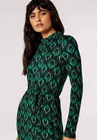 Abito a maniche lunghe verde e nero con motivo a foglie, caratterizzato da un colletto alto e una cintura legata in vita per una silhouette aderente.
