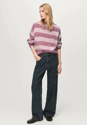 Mujer con cabello largo y rubio, vestida con un suéter de rayas rosa, jeans oscuros de pierna ancha, zapatos negros, con una mano en el bolsillo, mirando hacia la izquierda.