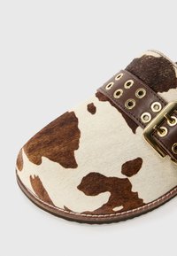 Leder-Slipper aus Rindsleder mit einem braun-weißen Muster, ausgestattet mit einem braunen Lederband, das goldene Ösen und eine Schnalle aufweist.