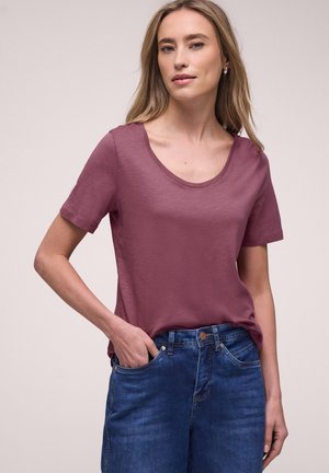 Donna con lunghi capelli biondi che indossa una maglietta a maniche corte color malva con scollatura a girocollo e jeans blu, che posa con una mano in tasca.