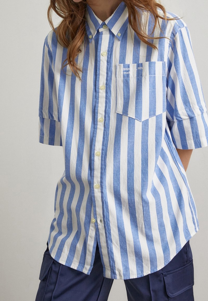 Camicia a maniche corte azzurro e bianco a righe, con taschino sul petto. Realizzata in un tessuto leggero, presenta bottoni gialli e una vestibilità comoda.