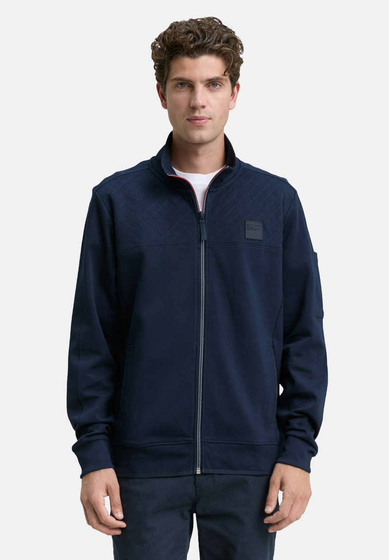 Jeune homme avec des cheveux bouclés portant une veste zippée bleu marine avec un haut de poitrine matelassé et une petite broderie, se tenant devant un fond uni.