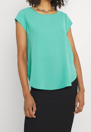 Femme portant un chemisier turquoise sans manches avec un col rond et une jupe noire, accessoirisée d'un collier chaîne argenté.