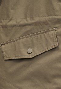 Gros plan sur un tissu kaki présentant une poche à rabat rectangulaire fermée par un seul bouton rond de couleur assortie.