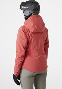 Rote isolierte Jacke mit taillierter Passform, verstellbarem Kapuzen und Frontreißverschlusstaschen. Getragen über beigen Hosen und ergänzt durch schwarze Handschuhe.