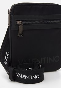 Svart kompakt crossbody-väska med framfickas dragkedja, metalldragkedjor med varumärkeslogotyp och justerbar rem med vit "VALENTINO"-text.