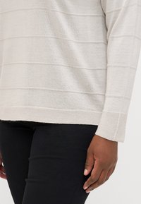 Vaaleanbeige pitkähihaisessa neulepaidassa on vaakatasoinen ribbitekstuuri ja suora helma, ja se on yhdistetty mustiin slim fit -housuihin.