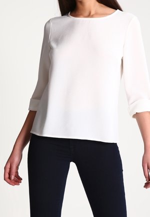 Kobieta ubrana w prostą białą bluzkę z długim rękawem z podwiniętymi mankietami i dopasowane ciemne spodnie, stojąca z lekko ugiętymi ramionami.