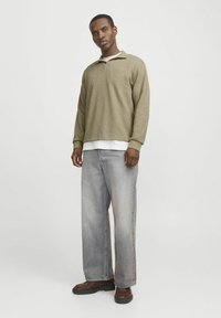 Olivegrünes Langarmoberteil mit Kragen, kombiniert mit hellgrauen weiten Jeans. Braune Schuhe runden das Outfit ab. Glatte Textur, lockere Passform.